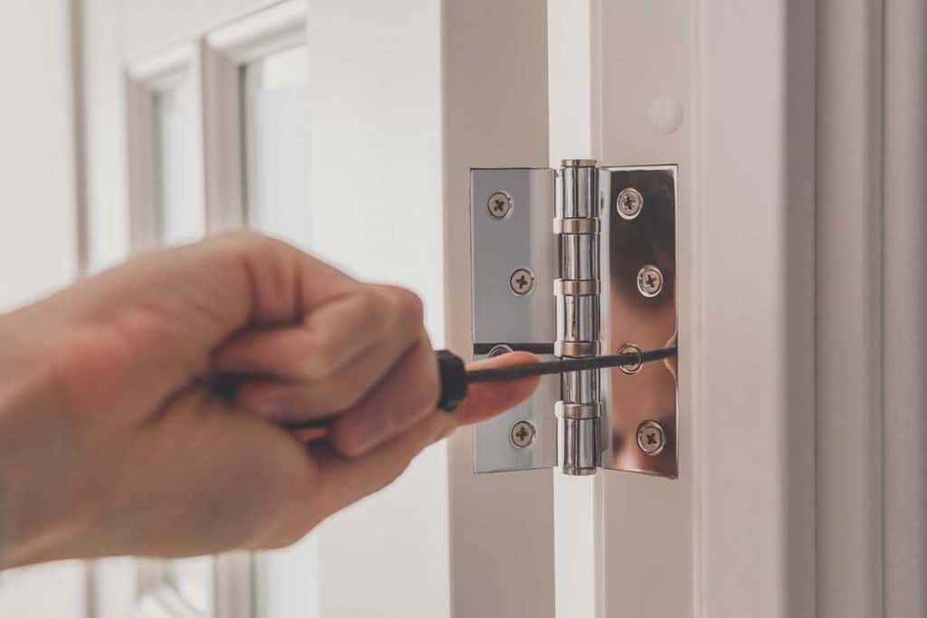 Man using screwdriver to remove screw from stainless door hinges on a white door