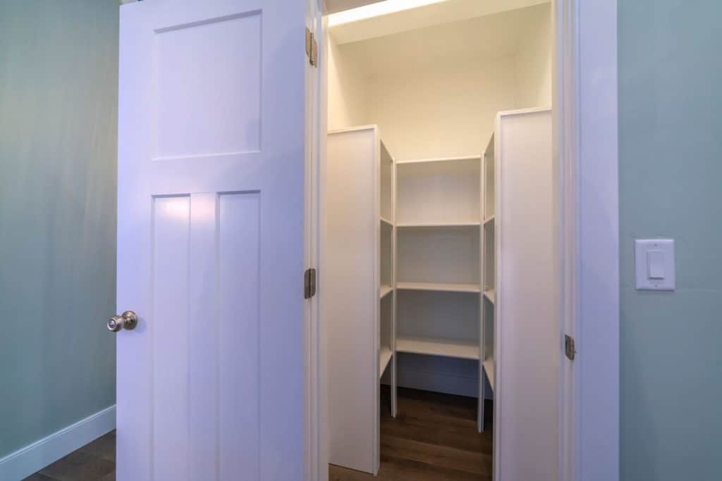 Looking into the empty interior of a walk-in pantry with rows of fitted white wooden shelves on and lights on, How Small Can A Pantry Door Be?