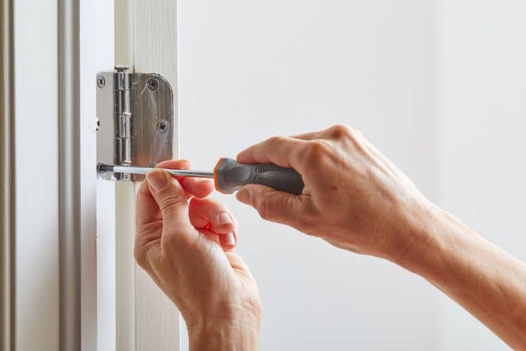 A carpenter installing a stainless steel door hinge using a screw driver, How To Fix Squeaky Door Hinges Without WD-40 [Would Vaseline Work?]