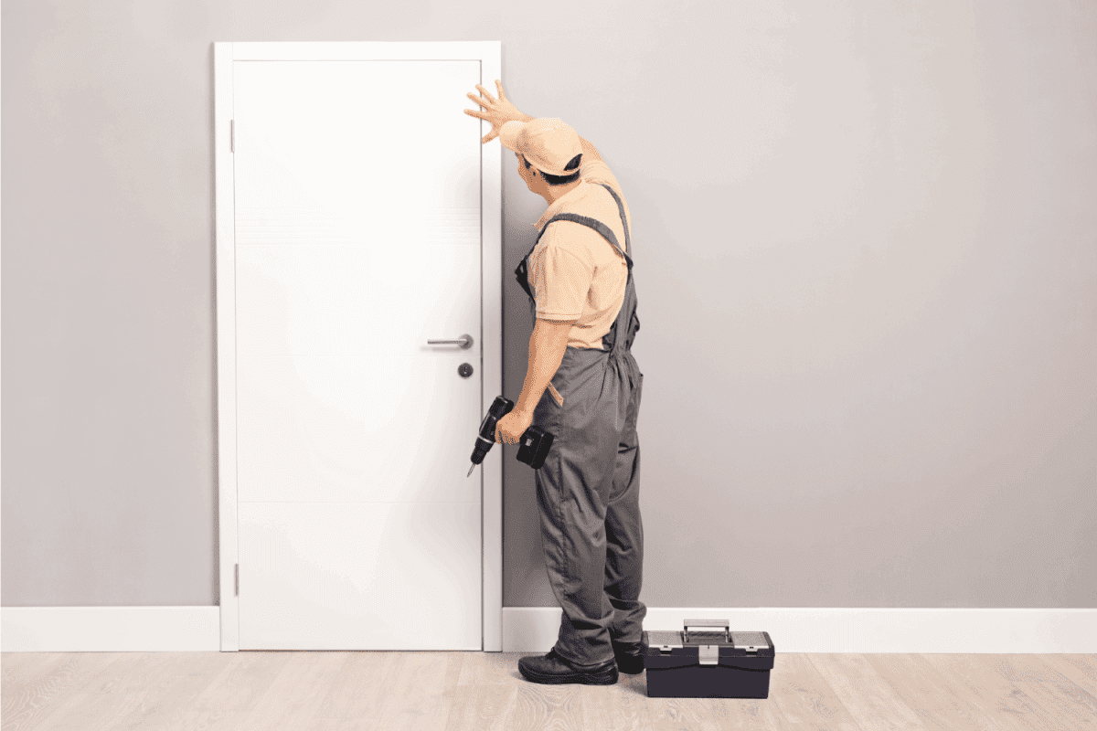 Young handyman installing a white door with an electric hand drill in a room