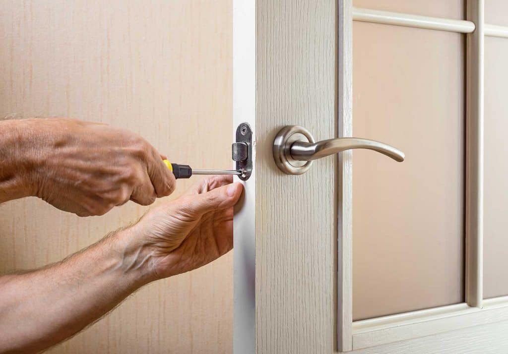 A man is mounting the protection strike of the deadbolt on a glass door with a modern curved style nickel handle using a screwdriver